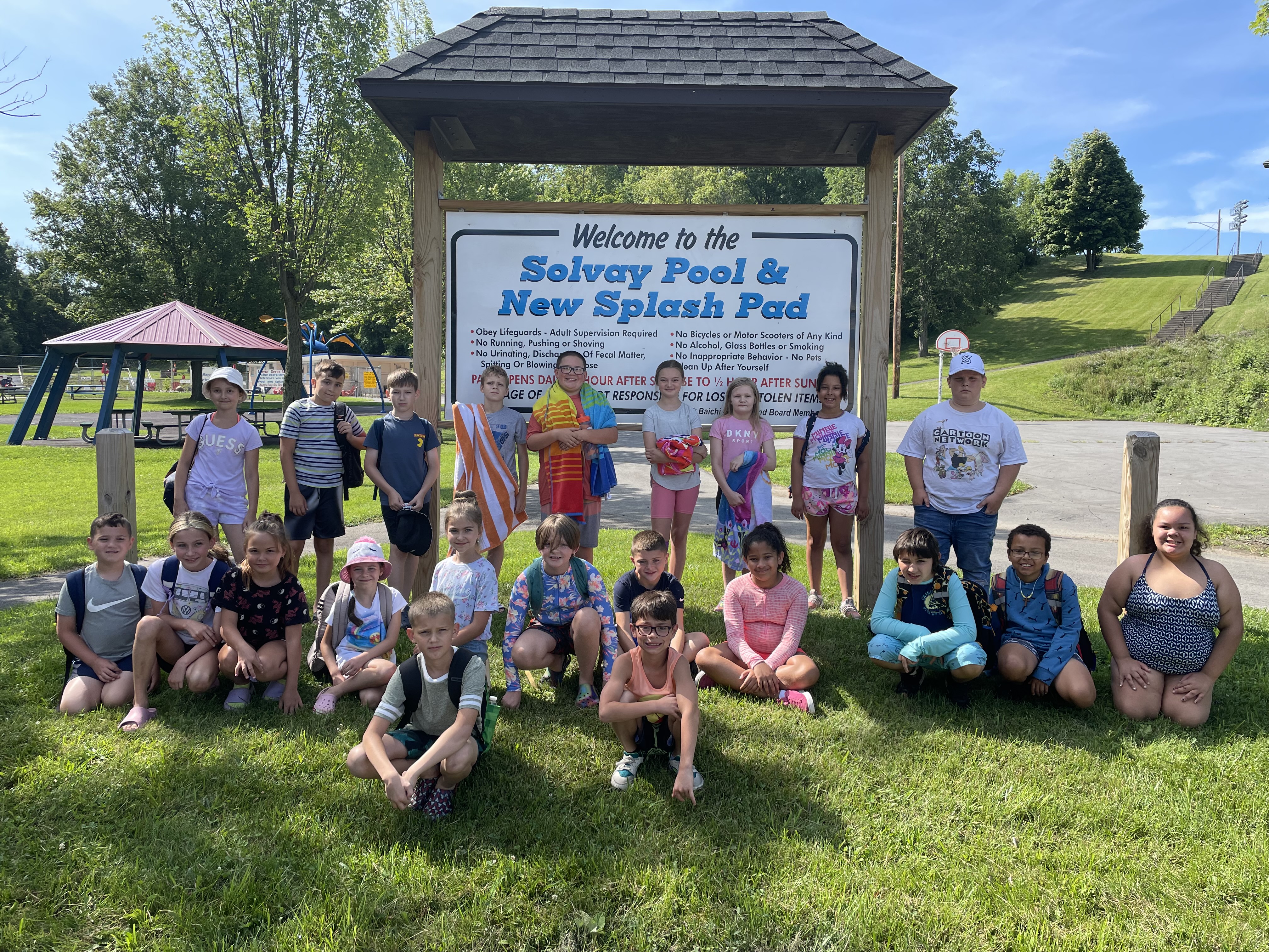 students sit on grass near splash pad sign